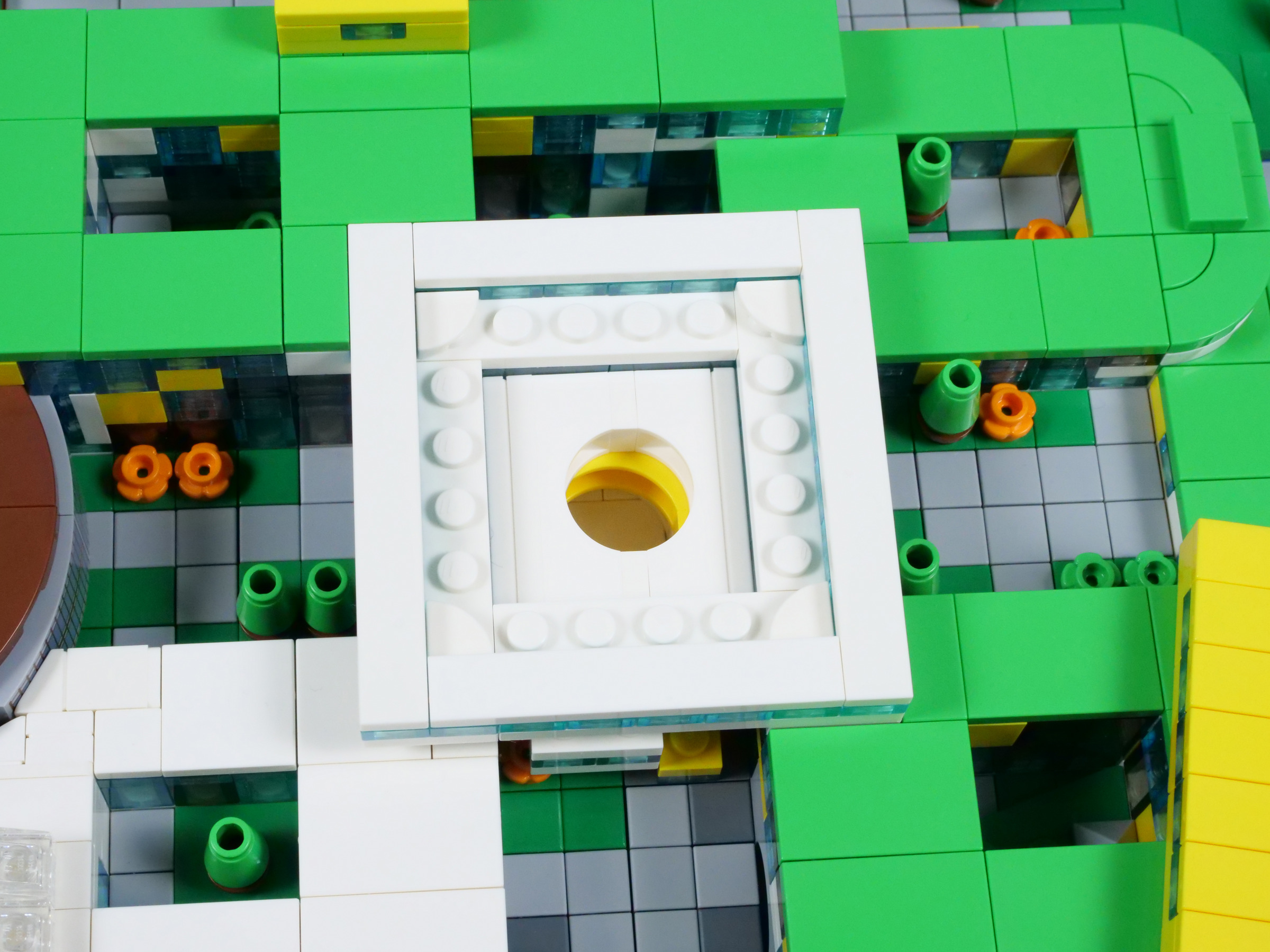Photographie Brickset du Campus vu du dessus : toit de l’atrium retiré, cadre carré blanc et ouverture ronde blanche et jaune au centre, toitures vertes, cours grises, arbres et fleurs autour.