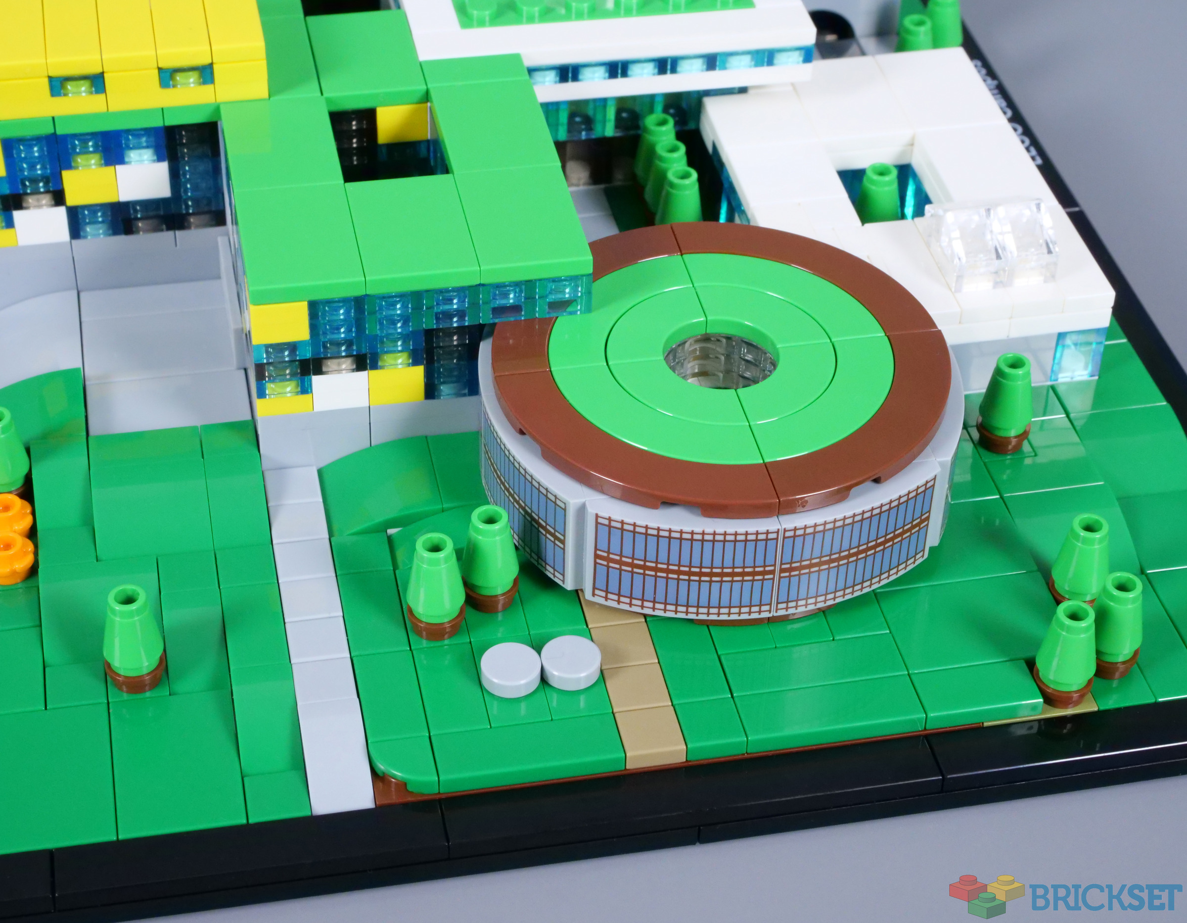Photographie Brickset de la cafétéria ronde du Campus : toit vert annulaire à trou central, quatre quarts intérieurs visibles, bordure brune, façade courbe à motifs de fenêtres, allée et petits arbres sur le socle.