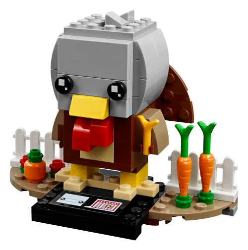 Dinde LEGO BrickHeadz à tête grise, bec jaune, appendice rouge et corps brun, sur un socle noir entouré de clôtures blanches, de carottes et d’une citrouille.